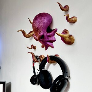 May include: A purple and gold octopus wall mount with eight tentacles, used to hang headphones. The octopus is 3D printed and has a textured surface.