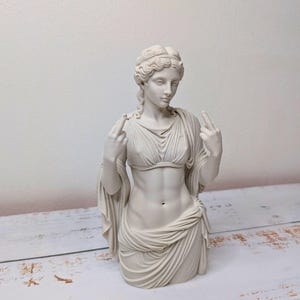 May include: A matte off-white bust sculpture of a classical figure with wavy hair, wearing a draped garment that leaves the midriff exposed. The figure is depicted with both arms raised, extending the middle finger on each hand toward the viewer.