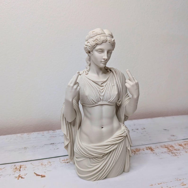 May include: A matte off-white bust sculpture of a classical figure with wavy hair, wearing a draped garment that leaves the midriff exposed. The figure is depicted with both arms raised, extending the middle finger on each hand toward the viewer.