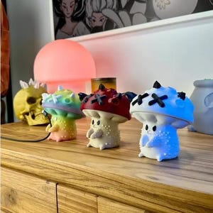 May include: Four colorful mushroom-shaped figurines with painted details. The figurines are made of a white material and have different colored accents. The figurines are arranged in a row on a wooden surface.