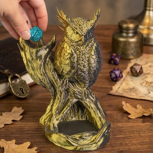 May include: A detailed owl figurine in gold and black, perched on a tree branch, designed to hold dice. A hand is placing a teal, multi-sided die on the branch. Other dice and a map are in the background. The owl has intricate feather detailing.