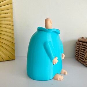 May include: A blue ceramic figurine of a person with a rounded body and no arms. The figurine has a small, rounded head with a light brown ear. The figurine is sitting on a white surface.