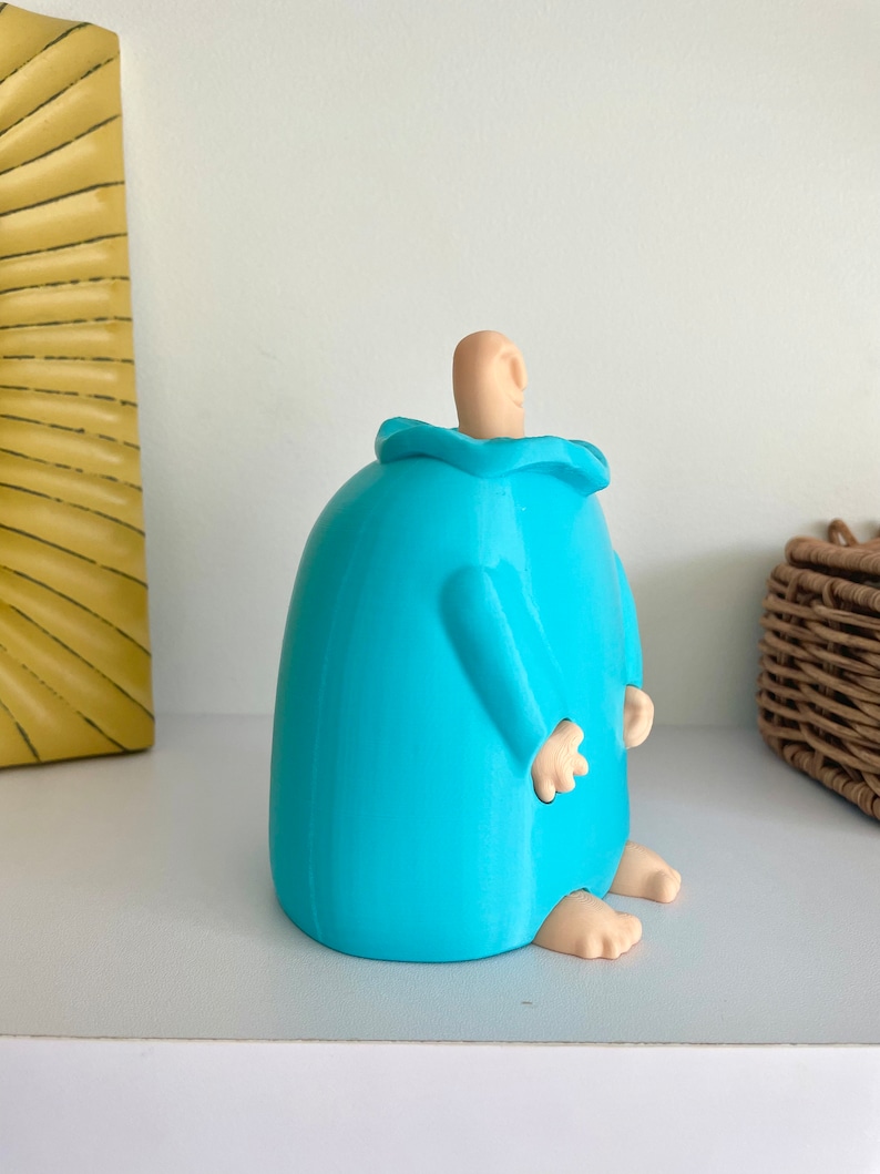 May include: A blue ceramic figurine of a person with a rounded body and no arms. The figurine has a small, rounded head with a light brown ear. The figurine is sitting on a white surface.