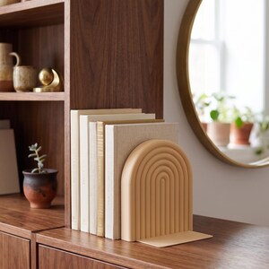 Op de afbeelding: Een houten boekenkast met decoratieve items en een set boeken die worden vastgehouden door een beige boogvormige boekensteun. De boeken hebben cr&egrave;me- en gouden covers. Een ronde gouden spiegel hangt aan de muur.