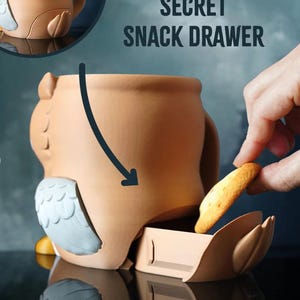 May include: A ceramic owl-shaped container with a secret snack drawer. The container is tan with gray and white accents. The text "SECRET SNACK DRAWER" is visible. A hand is placing a cracker into the drawer.