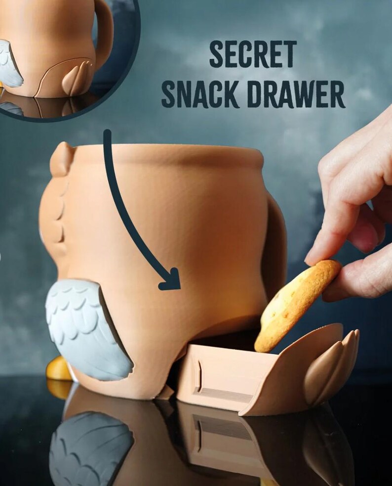 May include: A ceramic owl-shaped container with a secret snack drawer. The container is tan with gray and white accents. The text "SECRET SNACK DRAWER" is visible. A hand is placing a cracker into the drawer.