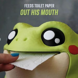 May include: A whimsical, lime green frog-shaped toilet paper holder. The frog has large, black and white eyes, a red cheek, and a smiling mouth dispensing toilet paper. Text above reads "FEEDS TOILET PAPER OUT HIS MOUTH."