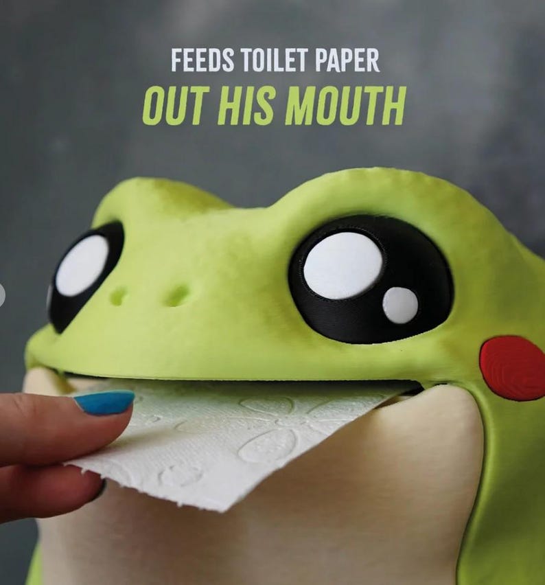 May include: A whimsical, lime green frog-shaped toilet paper holder. The frog has large, black and white eyes, a red cheek, and a smiling mouth dispensing toilet paper. Text above reads "FEEDS TOILET PAPER OUT HIS MOUTH."