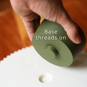 Puede incluir: Un objeto cil&iacute;ndrico verde oliva con una base roscada, colocado sobre una superficie blanca con un agujero circular. El texto "Base threads on" es visible. La imagen se centra en el dise&ntilde;o del objeto.