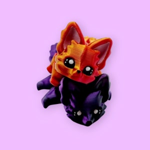Puede incluir: Una figurita impresa en 3D de dos zorros apilados uno encima del otro. El zorro de arriba es naranja y rojo con ojos negros. El zorro de abajo es morado con ojos negros.