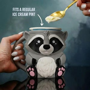 May include: A gray raccoon-shaped mug with black and white facial features, pink paw pads, and a white belly. The mug is designed to fit a regular ice cream pint. A golden spoon scoops ice cream from above.