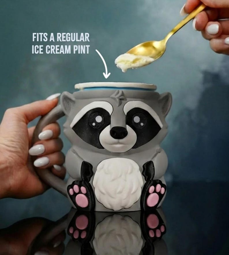 May include: A gray raccoon-shaped mug with black and white facial features, pink paw pads, and a white belly. The mug is designed to fit a regular ice cream pint. A golden spoon scoops ice cream from above.
