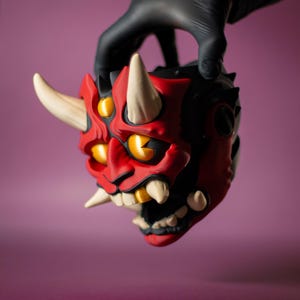 May include: A red and black Oni mask with white horns and yellow eyes. The mask has sharp teeth and is held by a gloved hand. The mask is a decorative item, possibly for cosplay or display.