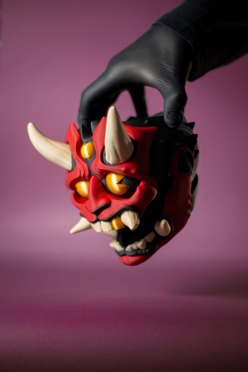 May include: A red and black Oni mask with white horns and yellow eyes. The mask has sharp teeth and is held by a gloved hand. The mask is a decorative item, possibly for cosplay or display.