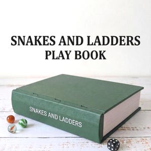 May include: A green "Snakes and Ladders Play Book" with white text. The book is closed and has a dark green cover. Three marbles and a black die with white dots are next to the book.