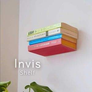 Op de afbeelding: Een wandplank gemaakt van een stapel boeken in verschillende kleuren, waaronder rood, blauw en geel. De boeken, getiteld "Alchemy", "You are the Placebo", "Your Next Five Moves" en "Home Ownership", zijn zo gerangschikt dat ze een zwevend schap creëren. De tekst "Invis Shelf" wordt hieronder weergegeven.