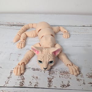 May include: A nude-colored, articulated Sphynx cat doll with a detailed face, large ears, and articulated limbs. The doll is lying on a light-colored surface, with its limbs extended. The cat's eyes are a dark color.