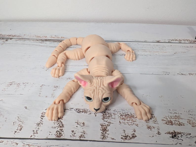 May include: A nude-colored, articulated Sphynx cat doll with a detailed face, large ears, and articulated limbs. The doll is lying on a light-colored surface, with its limbs extended. The cat's eyes are a dark color.