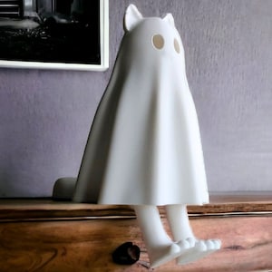 K&ouml;nnte beinhalten: Eine wei&szlig;e Keramikfigur in Form einer Katze im Geisterkost&uuml;m. Die Figur hat einen drapierten K&ouml;rper, Katzenohren und Augenl&ouml;cher. Die Figur sitzt mit h&auml;ngenden Beinen. Die Figur ist ein dekorativer Gegenstand.