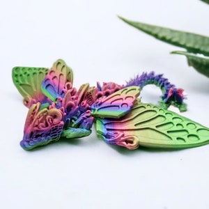 May include: A small, multi-coloured, 3D printed dragon figurine with wings that resemble butterfly wings. The dragon is lying on its side with its head turned to the left. The wings are a mix of green, pink, and purple.