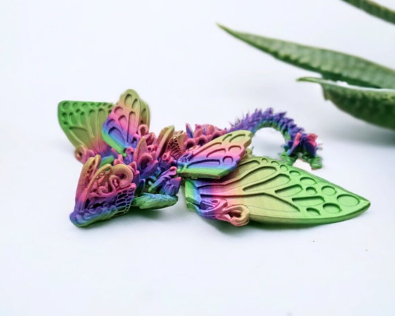 May include: A small, multi-coloured, 3D printed dragon figurine with wings that resemble butterfly wings. The dragon is lying on its side with its head turned to the left. The wings are a mix of green, pink, and purple.