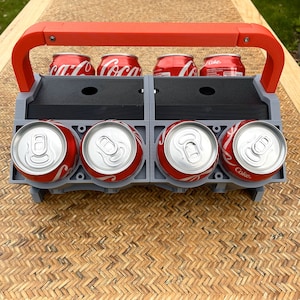 May include: A gray plastic six-pack can holder with a red handle. The holder is filled with six aluminum cans of soda. The cans have red labels with the word "Coke" on them.