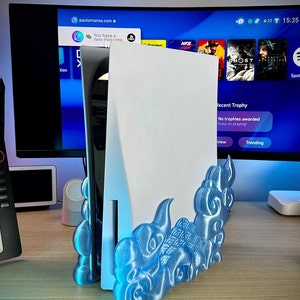 May include: A white PlayStation 5 console with a blue and white 3D printed stand shaped like clouds. The console is sitting on a wooden table.