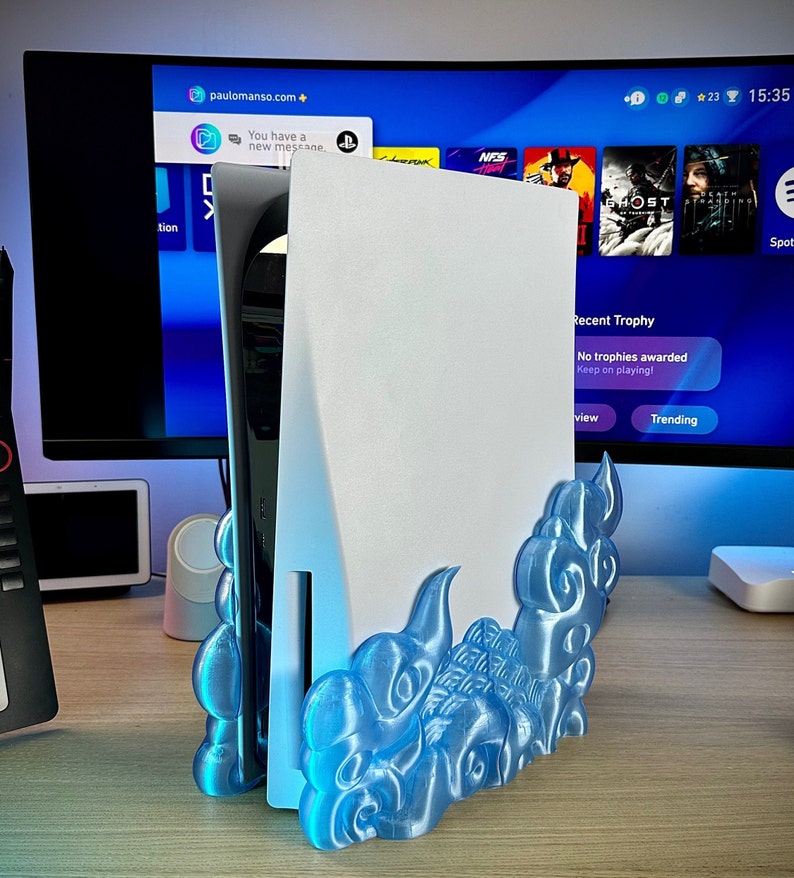 May include: A white PlayStation 5 console with a blue and white 3D printed stand shaped like clouds. The console is sitting on a wooden table.