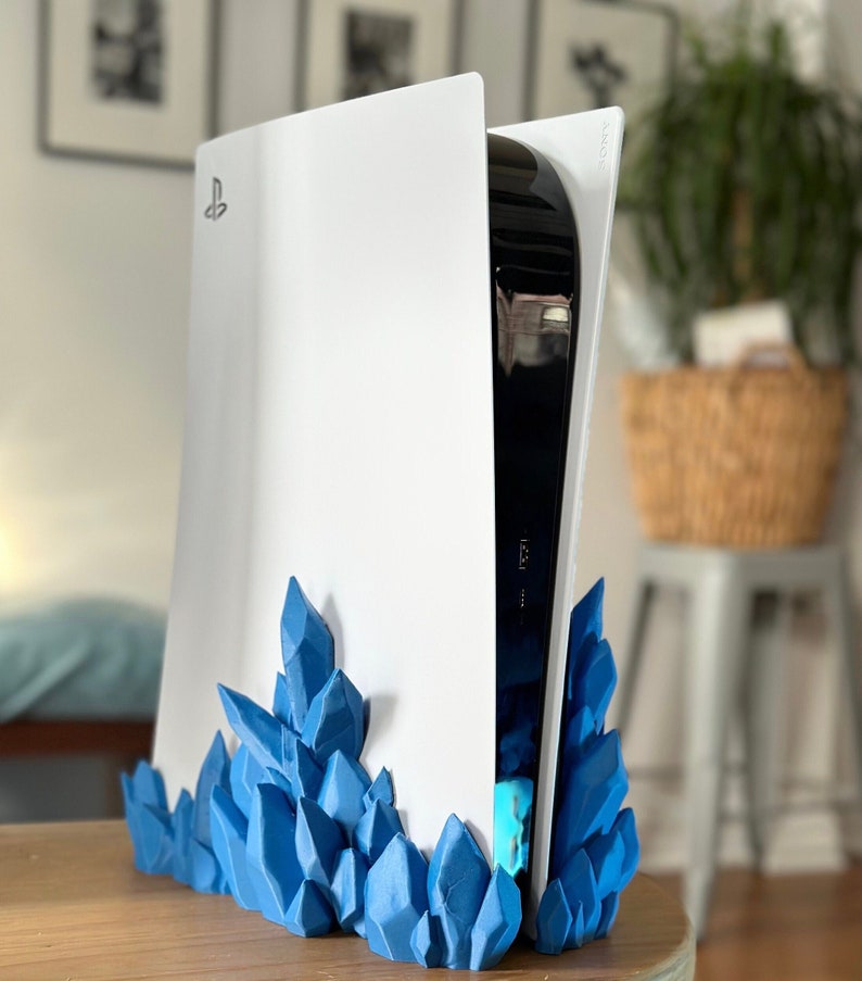 May include: A white PlayStation 5 console with a blue crystal-shaped stand. The console is standing upright and the stand is at the bottom of the console.