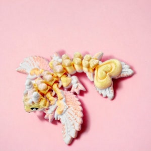 May include: A small, yellow and white, 3D printed dragon figurine with wings and a heart-shaped tail. The dragon is posed with its wings spread and its head tilted to the side.