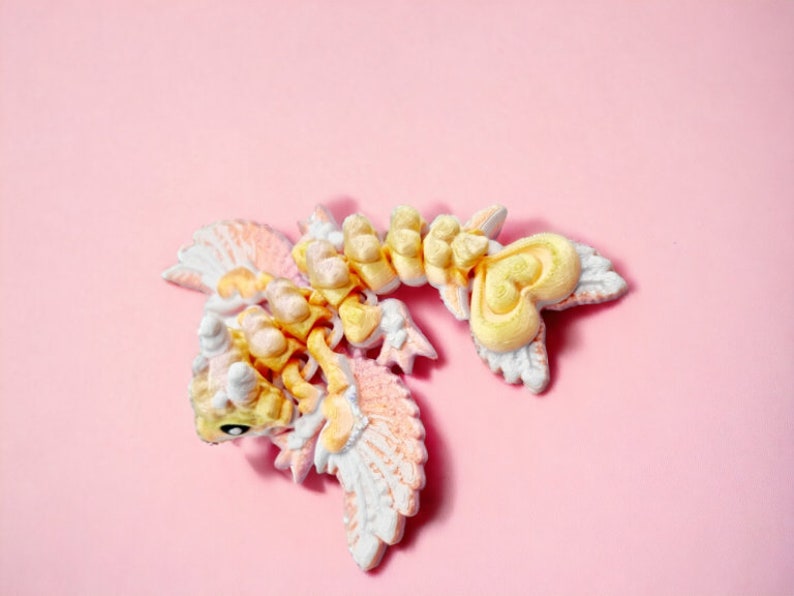 May include: A small, yellow and white, 3D printed dragon figurine with wings and a heart-shaped tail. The dragon is posed with its wings spread and its head tilted to the side.