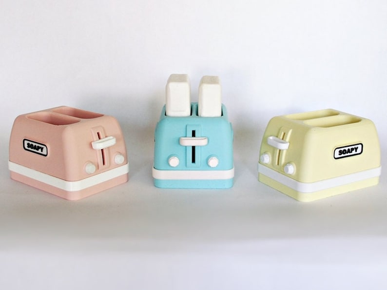K&ouml;nnte beinhalten: Drei Miniatur-Toaster in Pastellfarben: rosa, blau und gelb. Jeder Toaster hat einen Schlitz f&uuml;r zwei Brotscheiben und das Wort "SOAPY" ist auf der Vorderseite aufgedruckt.