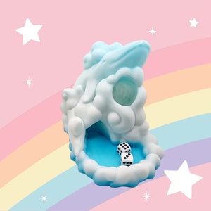 Puede incluir: Un original cubilete en forma de nube, en tonos azul y blanco, con dos dados en su interior. El cubilete se encuentra sobre un fondo pastel con un arco&iacute;ris y estrellas, creando una est&eacute;tica l&uacute;dica y so&ntilde;adora.