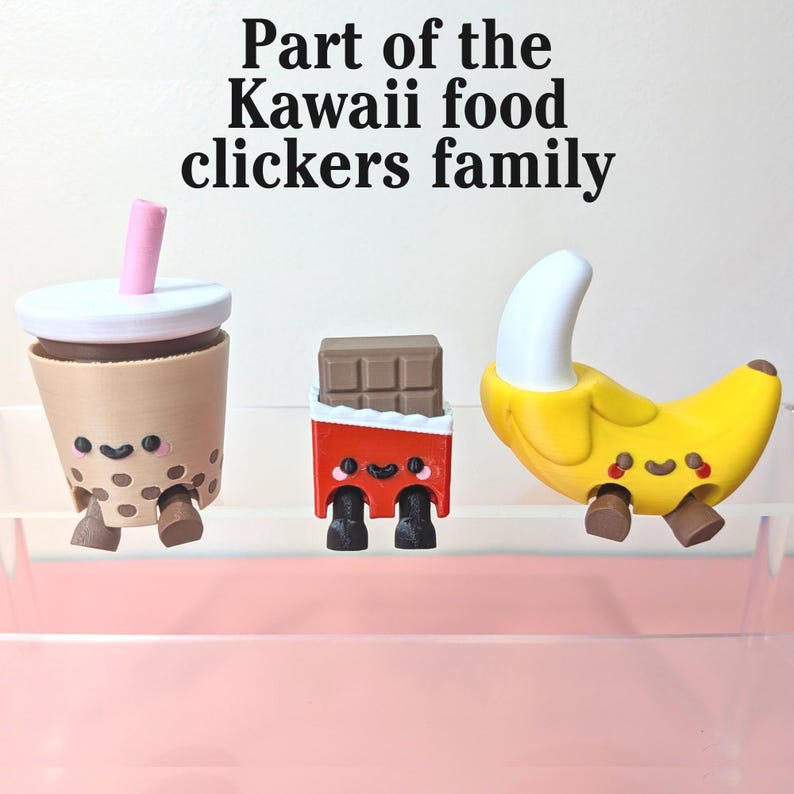 May include: Three Kawaii food clickers: a boba tea with a pink straw, a chocolate bar in red packaging, and a yellow banana. Each has a smiling face and small legs. Text reads "Part of the Kawaii food clickers family."