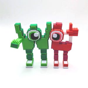 May include: Two pixelated figures, one green and one red, stand side-by-side. The green figure has a large eye and a hand-like appendage. The red figure has a similar eye and a hand making a gesture. Both figures have blocky legs and feet.