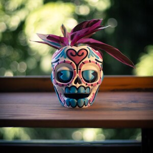 May include: A decorative skull-shaped planter with a colorful design, featuring pink, blue, and black accents. The planter holds a small plant with deep purple leaves. The planter sits on a wooden surface with a blurred green background.