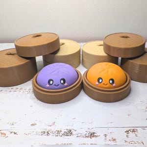 Op de afbeelding: Twee ronde, gladde, kleurrijke knoedelfiguren met lachende gezichten, &eacute;&eacute;n paars en &eacute;&eacute;n oranje, elk rustend in een bruine ronde container. Verschillende extra bruine, cilindrische containers met deksel zijn erachter geplaatst.