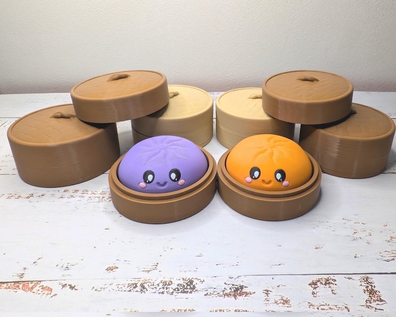 Op de afbeelding: Twee ronde, gladde, kleurrijke knoedelfiguren met lachende gezichten, &eacute;&eacute;n paars en &eacute;&eacute;n oranje, elk rustend in een bruine ronde container. Verschillende extra bruine, cilindrische containers met deksel zijn erachter geplaatst.