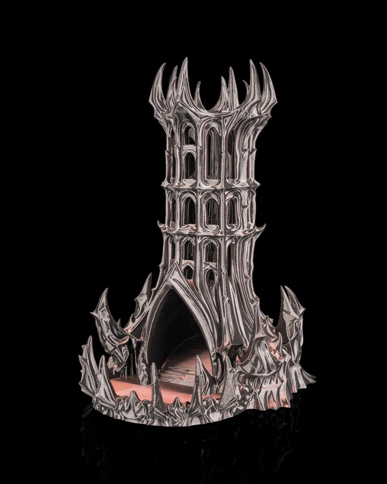 May include: A silver-colored, gothic-style tower sculpture with intricate architectural details. The tower features multiple levels with arched windows and a spiked crown. The base has a textured, fortress-like appearance, with a dark interior.