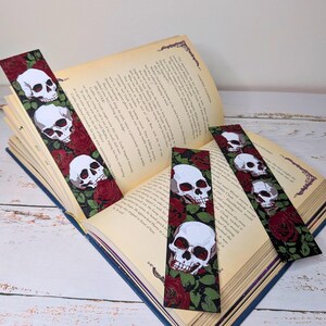 May include: Four decorative bookmarks with a skull and rose design. Each bookmark features white skulls with red accents against a black background with red roses and green leaves. The bookmarks are placed on an open book.