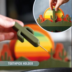 May include: A green cactus-shaped toothpick holder with a toothpick inserted. The image shows a desert-themed design with a sunset and cacti. The text "TOOTHPICK HOLDER" is visible.