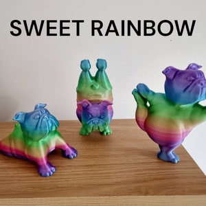 K&ouml;nnte beinhalten: Drei Regenbogenfarbene Bulldoggen-Figuren in verschiedenen Posen. Eine liegt, eine steht auf dem Kopf und die dritte macht Yoga. Der Text "SWEET RAINBOW" steht &uuml;ber den Figuren.