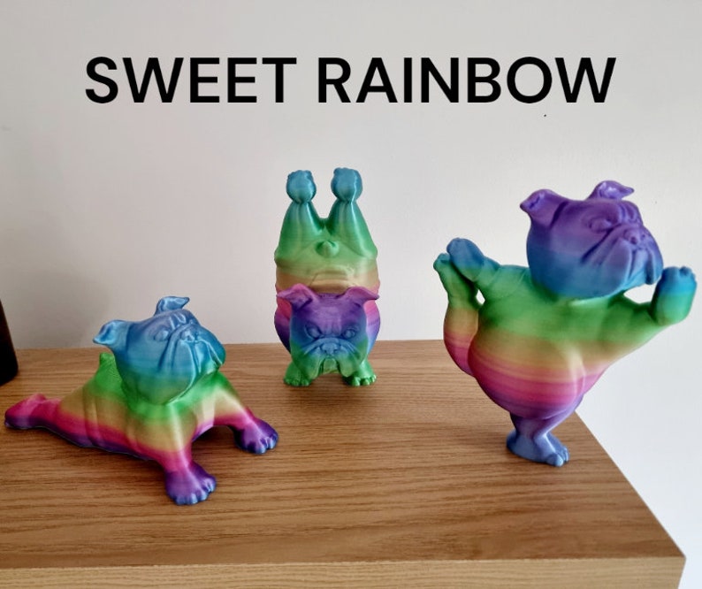 K&ouml;nnte beinhalten: Drei Regenbogenfarbene Bulldoggen-Figuren in verschiedenen Posen. Eine liegt, eine steht auf dem Kopf und die dritte macht Yoga. Der Text "SWEET RAINBOW" steht &uuml;ber den Figuren.