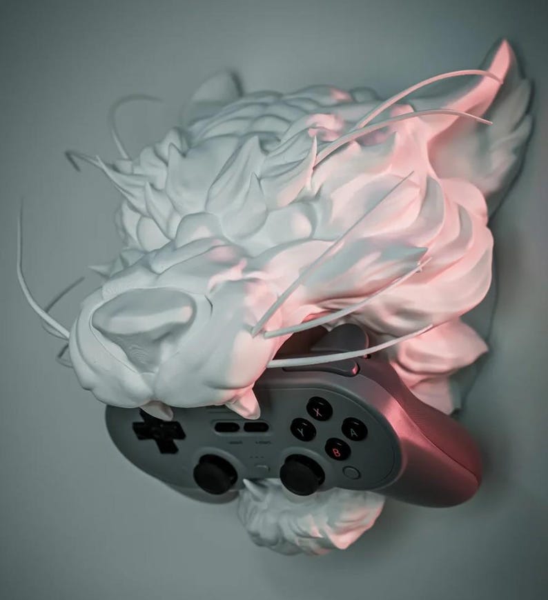 May include: A white, sculpted cat head with a gray video game controller. The cat head has a textured, cloud-like appearance. The controller has black buttons and a directional pad. The overall aesthetic is modern and artistic.