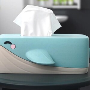 May include: A whimsical tissue box shaped like a blue whale. The box features a light blue body, a beige underside, and a tail. The whale has a smiling face with a pink cheek and a fin. White tissues are emerging from the top.