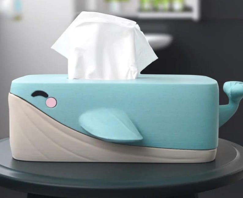 May include: A whimsical tissue box shaped like a blue whale. The box features a light blue body, a beige underside, and a tail. The whale has a smiling face with a pink cheek and a fin. White tissues are emerging from the top.