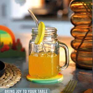 May include: A clear glass jar with a handle, filled with orange liquid and a lemon slice, sits on a rainbow-colored coaster. A glass straw is in the jar. The text "BRING JOY TO YOUR TABLE" is at the bottom of the image.