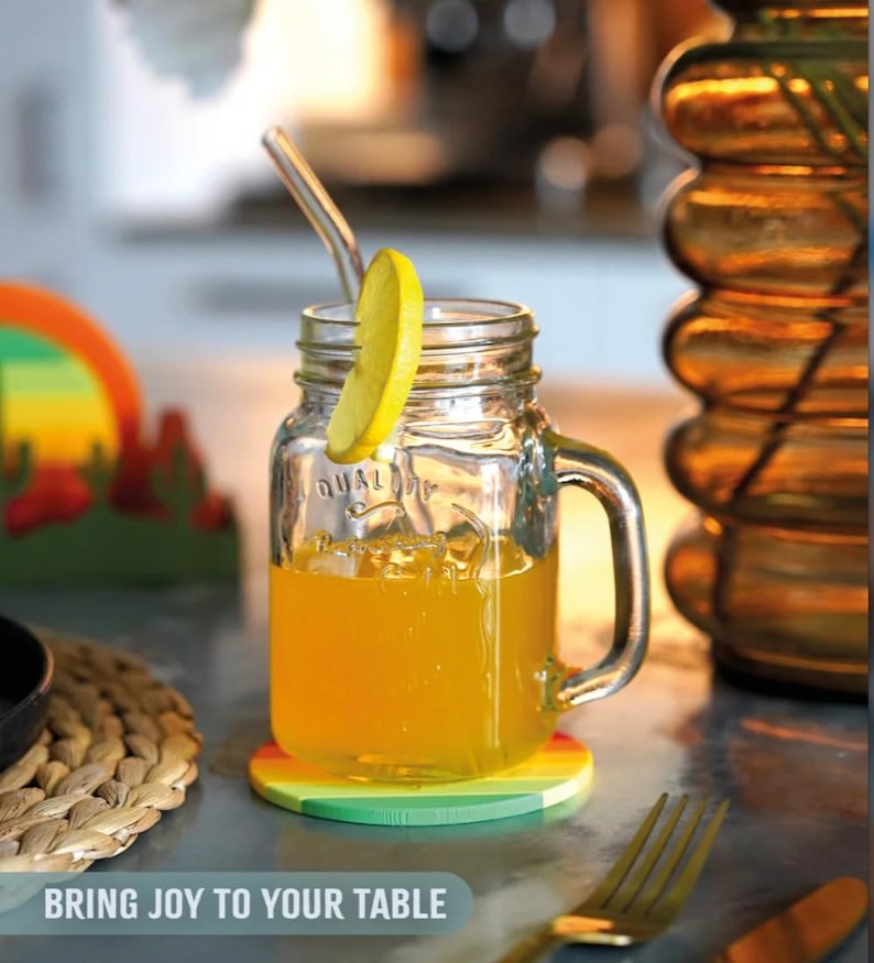 May include: A clear glass jar with a handle, filled with orange liquid and a lemon slice, sits on a rainbow-colored coaster. A glass straw is in the jar. The text "BRING JOY TO YOUR TABLE" is at the bottom of the image.
