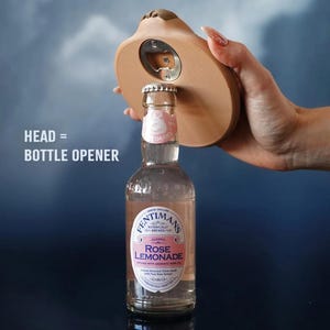 Op de afbeelding: Een novelty flesopener in de vorm van een hoofd, die een fles Rose Lemonade opent. De tekst op de afbeelding luidt "HEAD = BOTTLE OPENER". De fles is helder met een roze vloeistof erin.