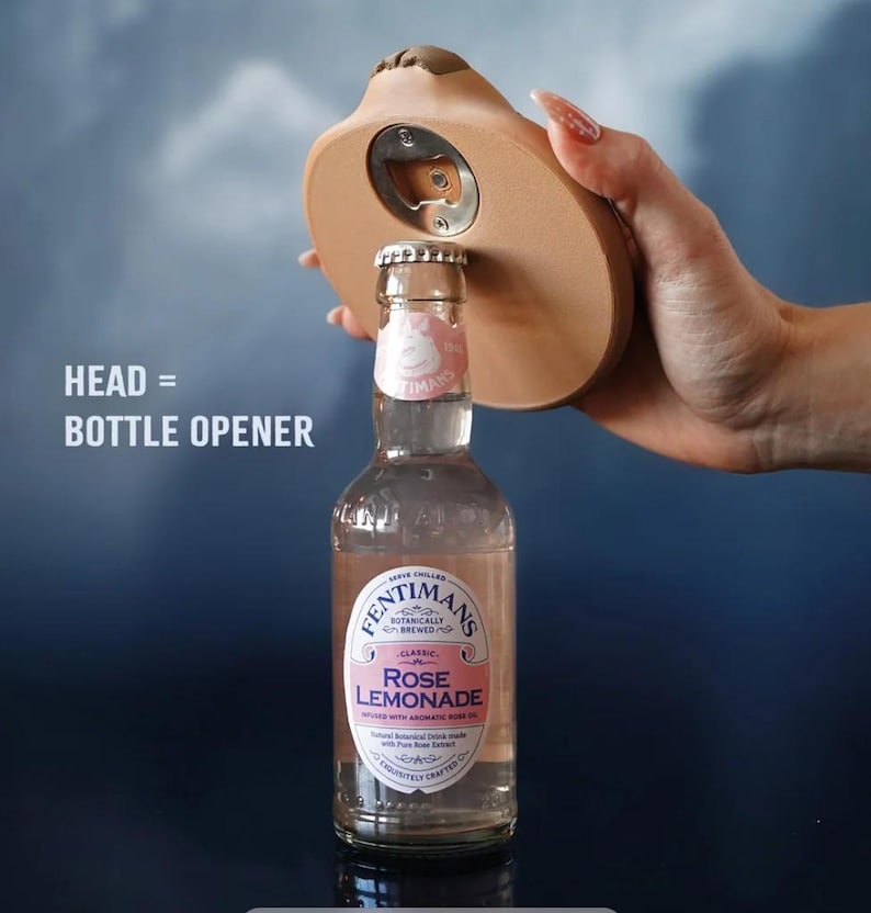Op de afbeelding: Een novelty flesopener in de vorm van een hoofd, die een fles Rose Lemonade opent. De tekst op de afbeelding luidt "HEAD = BOTTLE OPENER". De fles is helder met een roze vloeistof erin.